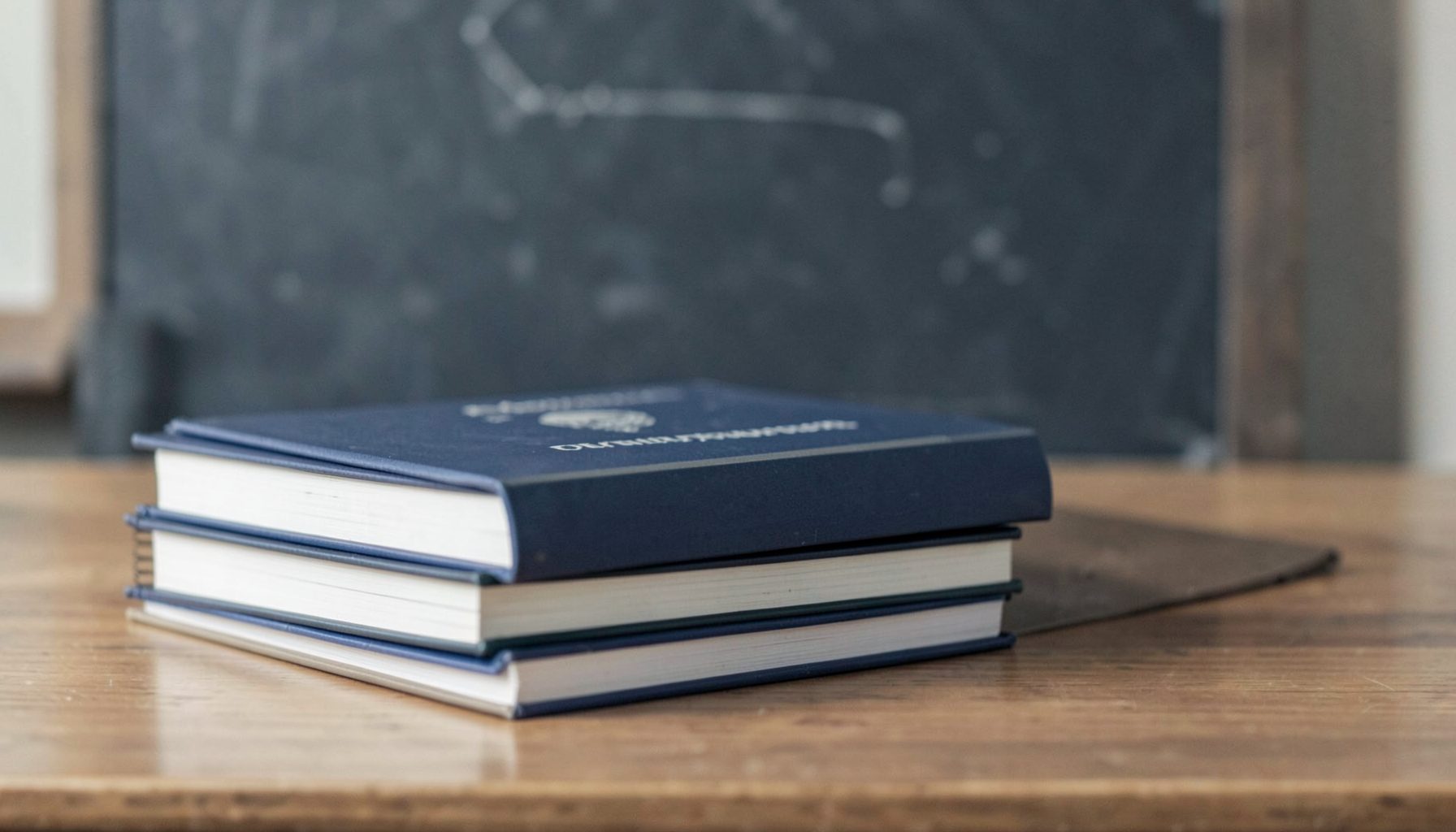 A stack of textbooks and curriculum binders on a wooden desk, with a chalkboard in the background. Muted, serious tones of navy blue, gray, and cream.